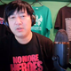 Suda51, wearing headphones and a No More Heroes shirt, sits in front of handful of tees on hangers.